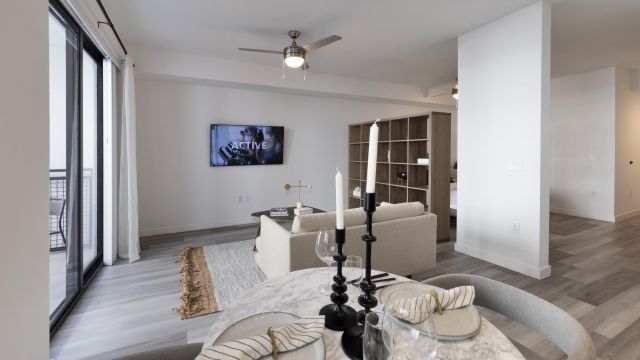 The Upland Modern living room with a round dining table, candlesticks, and a TV on the wall. Beige sofa and large window visible.