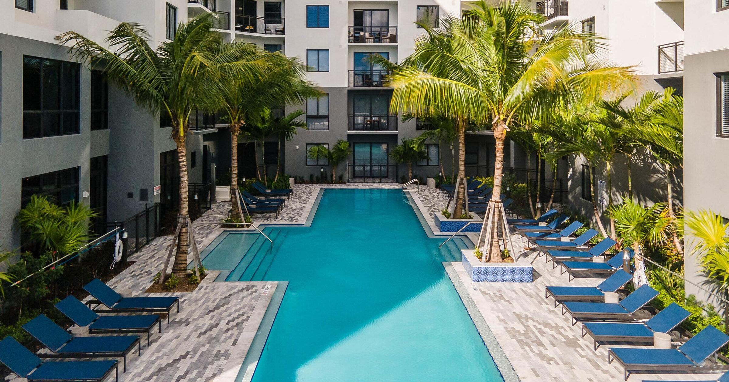 The Upland A rectangular pool surrounded by blue lounge chairs and palm trees in a courtyard of a tall building.