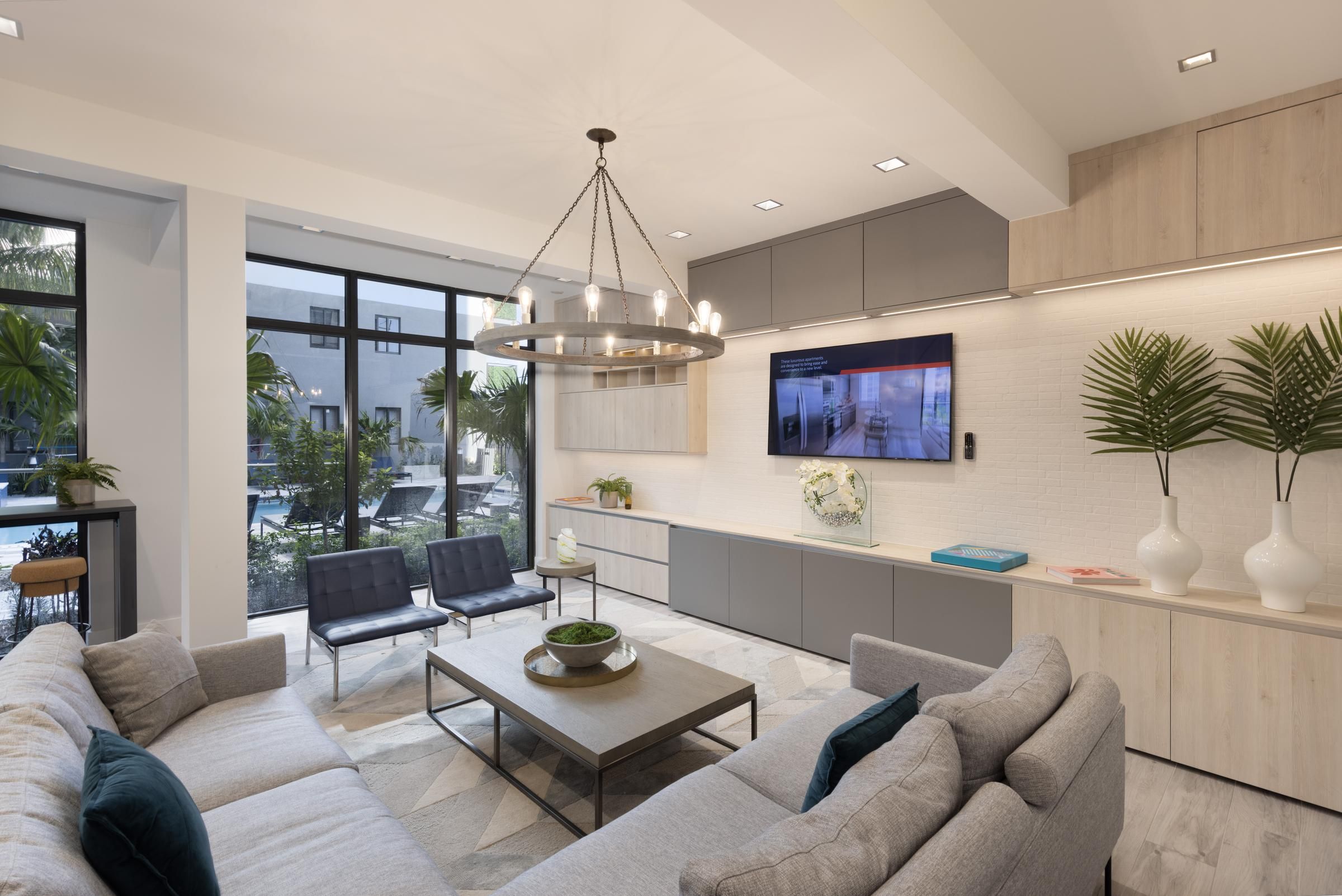 The Upland Modern living room with a gray sectional sofa, wall-mounted TV, and large windows overlooking a pool area.