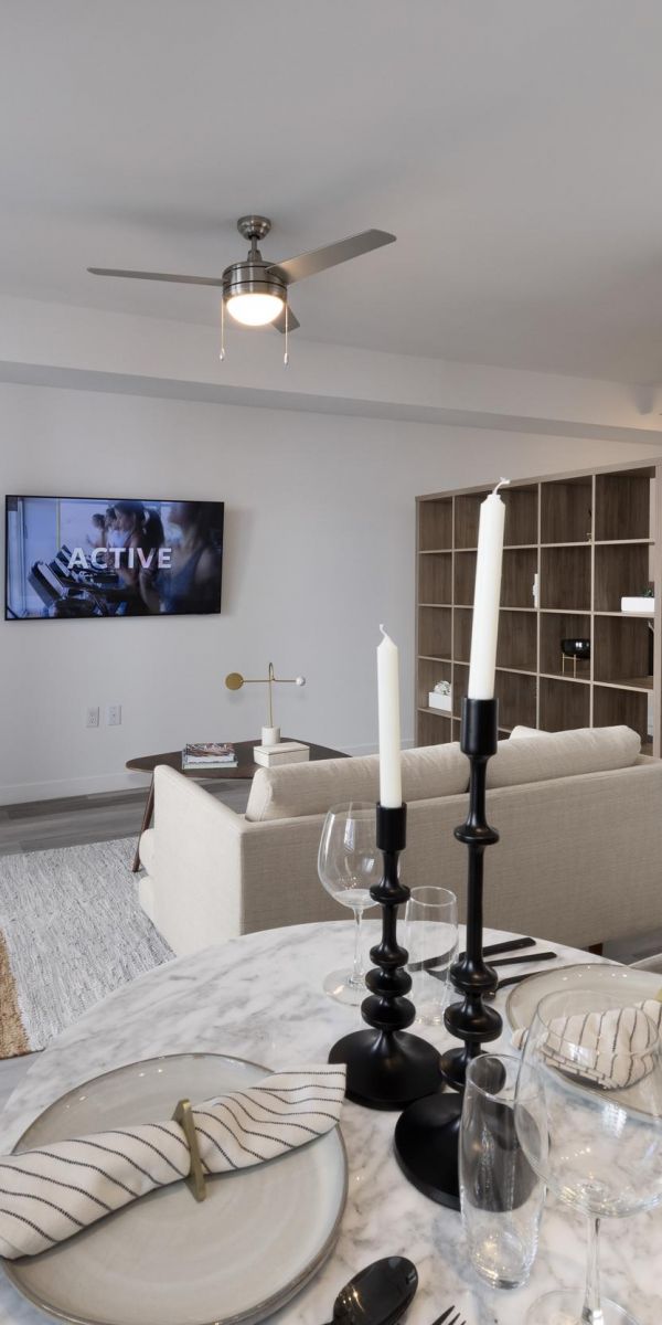 The Upland Modern living room with a round dining table, candlesticks, and a TV on the wall. Beige sofa and large window visible.