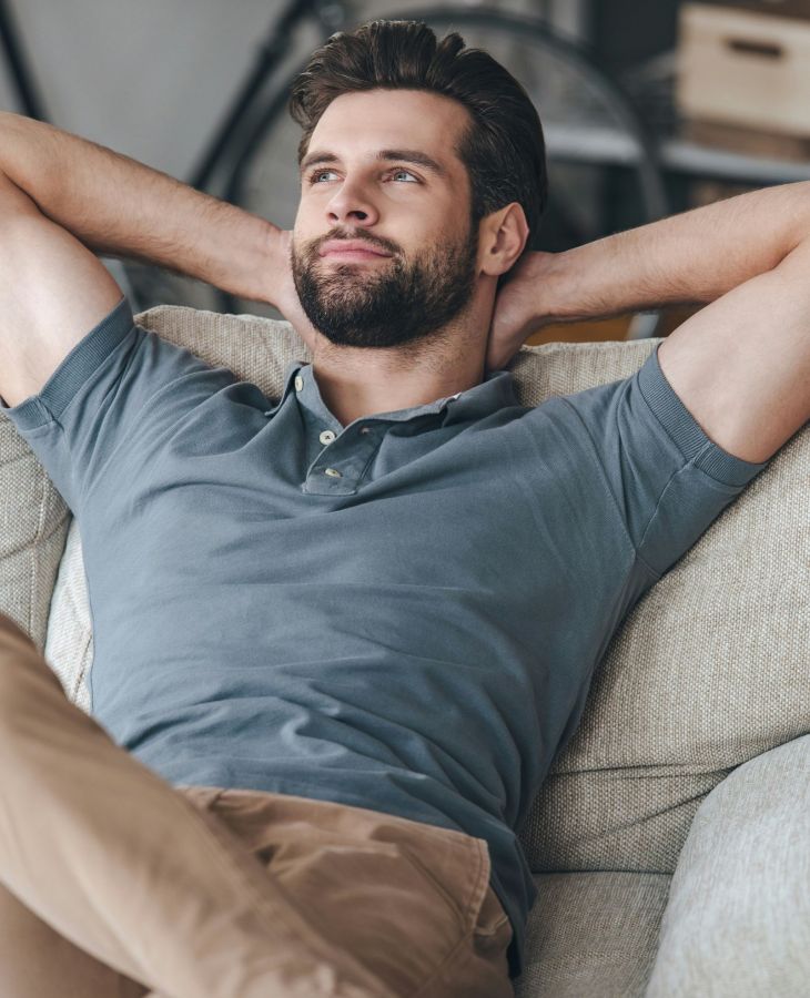 The Upland Man relaxing on a couch, wearing a gray polo shirt and brown pants, with hands behind head, looking content.