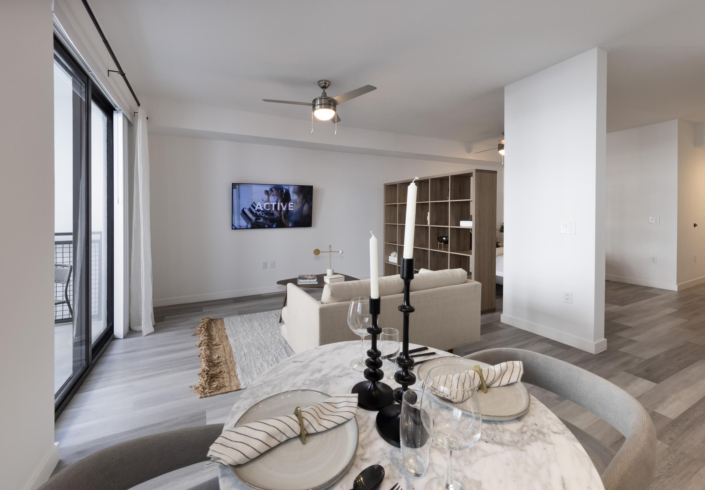 The Upland Modern living room with a round dining table, candlesticks, and a TV on the wall. Beige sofa and large window visible.