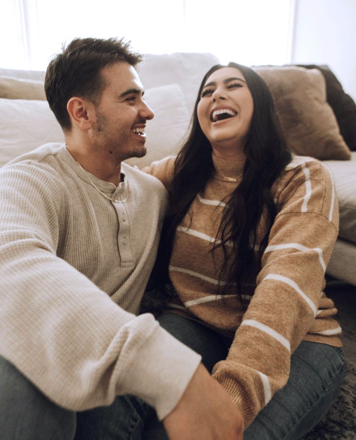 The Upland A couple sitting on the floor, laughing together, wearing casual sweaters and jeans in a cozy living room.