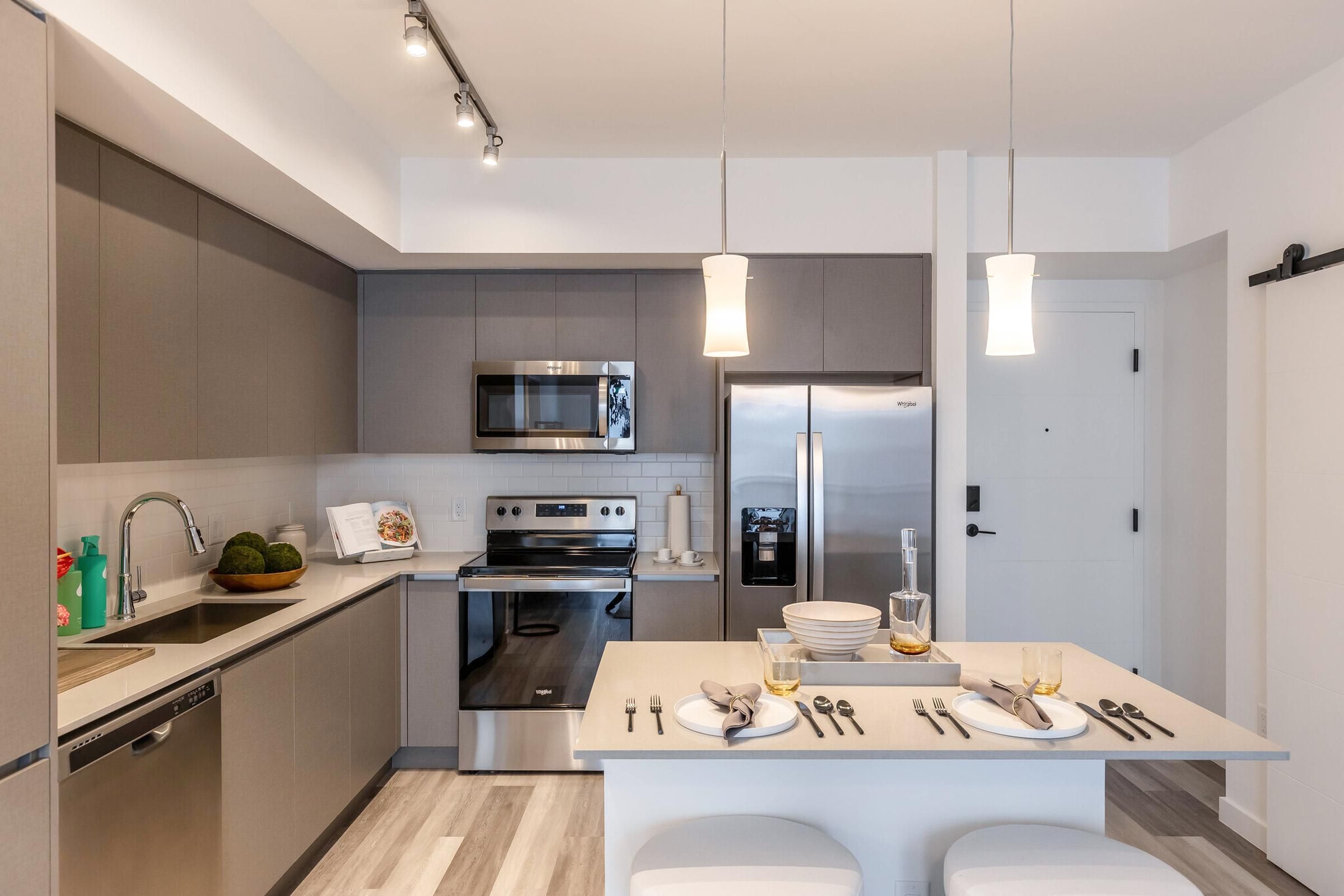 The Upland Modern kitchen with gray cabinets, stainless steel appliances, and a small island with two stools and tableware.